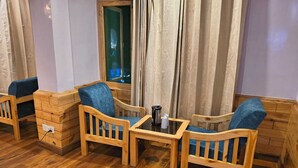 A cozy seating area with wooden chairs and a small table, a window with curtains, and a wooden floor. - Hotel Kasol Inn (Kasol)