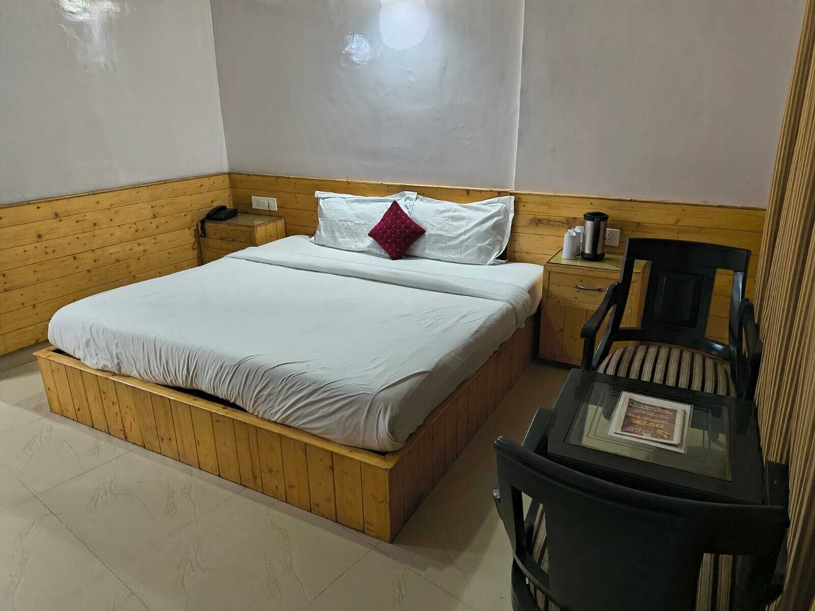 A bedroom with a wooden bed, a nightstand with a phone, a chair, and a table with a framed item.