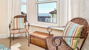 Una habitación con dos sillones de mimbre con cojines a rayas, un cofre de madera y vistas al mar a través de una ventana.