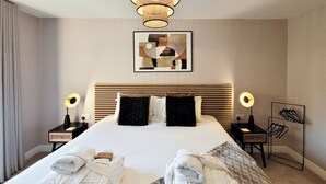 Et hotelværelse med en stor seng, to sengeborde med lamper, et indrammet abstrakt billede og et par håndklæder på sengen.