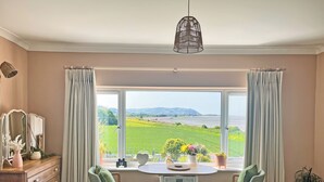 Et værelse med et stort vindue, der giver udsigt til et grønt landskab og bakker. Der er et træmøbel, et bord med stole og en hængende lampe.