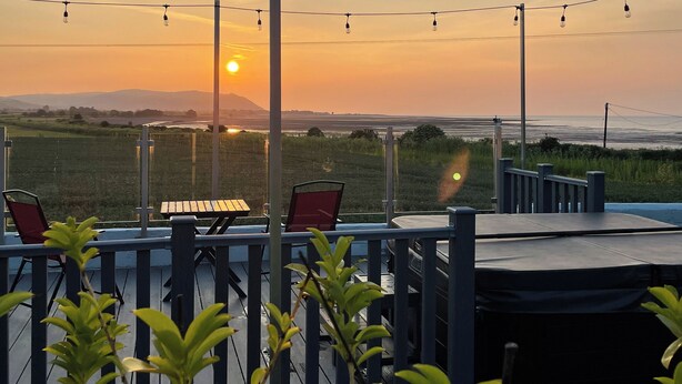 En udendørs siddeplads med udsigt til solnedgangen over havet, lysslinger og en træterrasse.