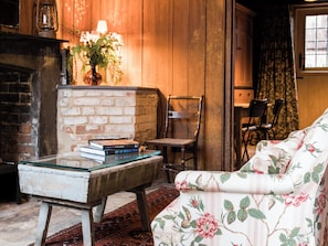 Una habitación acogedora con chimenea de ladrillo, una silla de madera, un sofá tapizado con motivos florales y una mesa con libros.