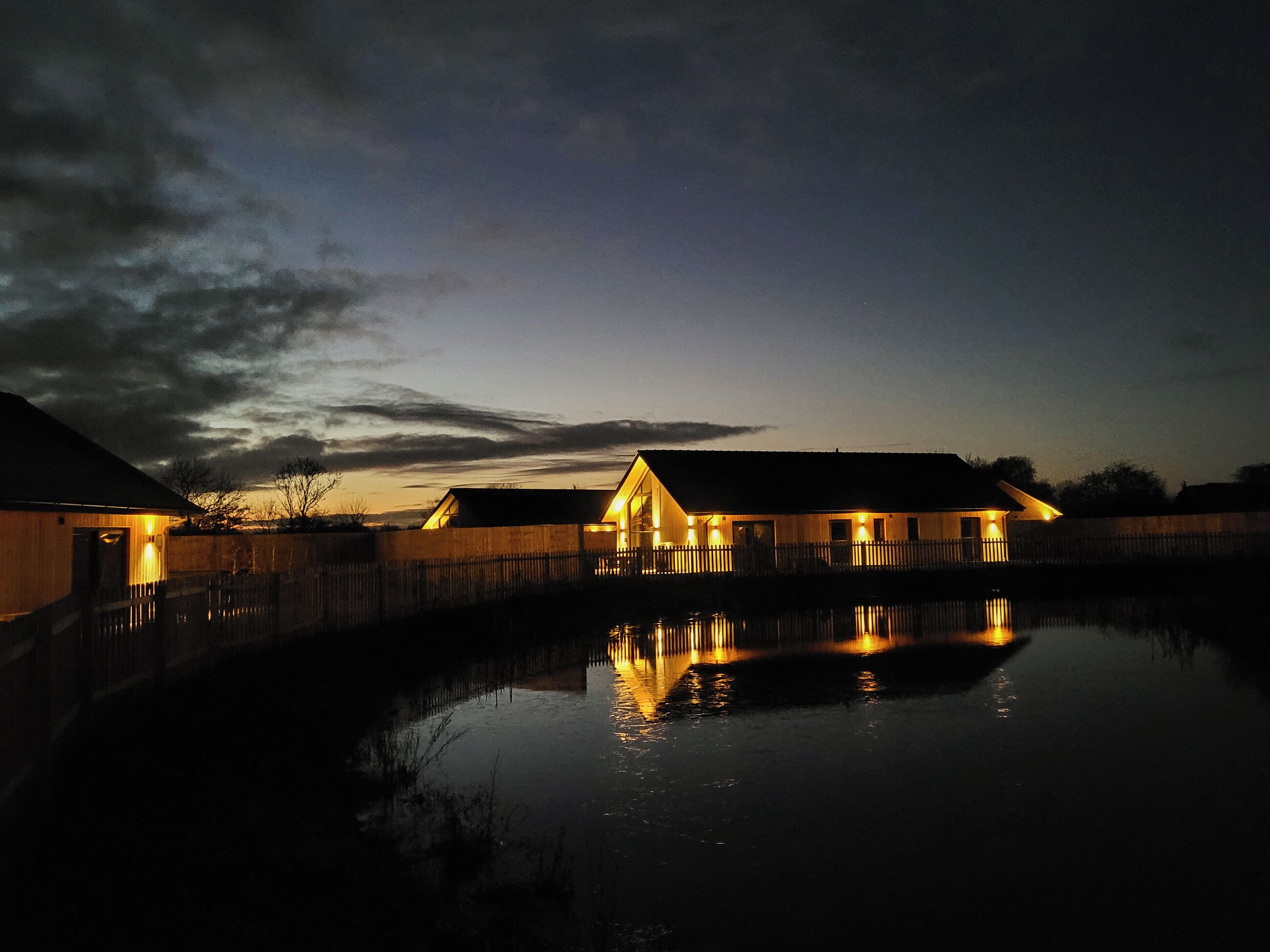 Une propriété située au bord de l’eau, avec des bâtiments illuminés dont les reflets se mirent dans l’eau, une clôture en bois et un ciel dégagé.