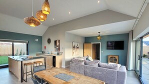 A modern kitchen with a central island, a dining area with a wooden table, and a living space with a grey sofa. - Bailgate Barn - UKC4646 (Dunholme, near Lincoln)