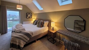 Een slaapkamer met een bed, nachtkastje, lamp en een spiegel. Er zijn dakramen en uitzicht op het omliggende landschap.