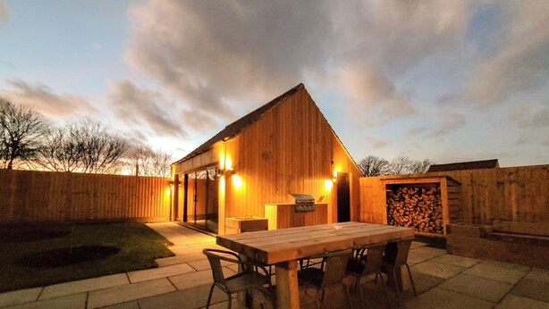 Une construction en bois avec un toit en pente, une terrasse avec une table et des chaises, et une pile de bois de chauffage.