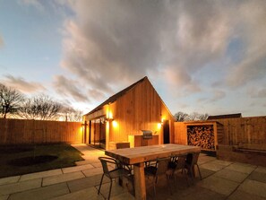 A wooden building with a sloped roof, a patio with a table and chairs, and a stack of firewood. - Lindum Barn - UKC4649 (Dunholme, near Lincoln)