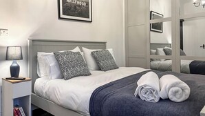 Ett välordnat sovrum med en säng, kuddar, ett sängbord med en lampa, en inramad tavla på väggen och en garderob med spegeldörrar.