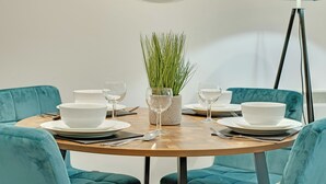 A dining table set with white plates, bowls, and cutlery, surrounded by teal upholstered chairs, with a green plant in a patterned vase and a floor lamp in the background. - All Saints Apartments 4A (Newcastle Upon Tyne)