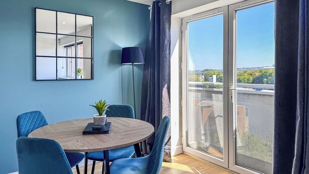 Ein modernes Zimmer mit einem runden Holztisch, blauen Stühlen, einem Blumentopf, einer Stehlampe und einer großen Schiebeglastür, die zu einem Balkon mit Blick auf die Stadt führt.