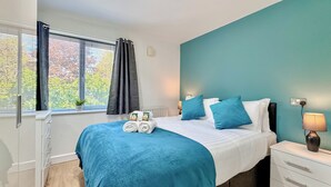Ein modernes Schlafzimmer mit einer Türkis-farbenen Akzentwand, einem ordentlich bezogenen Bett mit blau-weißer Bettwäsche, einem Nachttisch mit Lampe und einem Fenster mit Vorhängen.