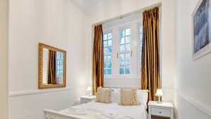 Een slaapkamer met een wit bed, twee nachtkastjes, een raam met gordijnen en een spiegel aan de muur.
