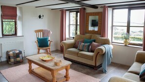 Ein gemütliches Wohnzimmer mit einem Holztisch, einem beigefarbenen Sofa mit vielen Kissen, einem Holzsesseln mit einem blauen Kissen und einem Fenster mit Blick auf Grünflächen.