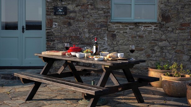 Ein rustikales Steinhaus mit einem Holztisch, gedeckt für eine Mahlzeit, einer Weinflasche und einem Schild mit der Aufschrift "DOOR COTTAGE".