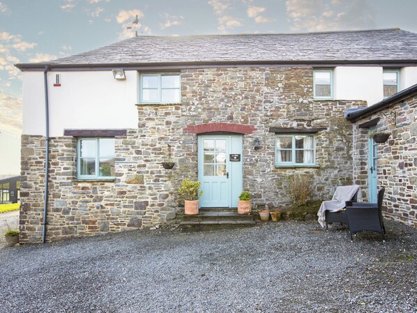 A stone building with a blue door, two windows, and a small porch with a chair. - Jasmine Cottage - UKC4694 (Woodford, near Bude)