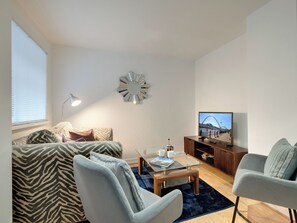 Ein modernes Wohnzimmer mit einer Couch, Sesseln, einem Couchtisch, einem Fernseher und einem dekorativen Spiegel.