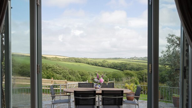 A patio with a table and chairs, a vase with flowers, and a view of green hills. - The Stables - UKC4696 (Woodford, near Bude)