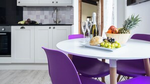 Une cuisine moderne avec des meubles blancs, un plan de travail noir et un coin repas comprenant une table blanche et des chaises violettes. Sur la table, on trouve deux verres à vin, une miche de pain et un saladier de fruits.