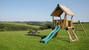 Une structure de jeux en bois, avec un toboggan et des balançoires, installée dans un champ d’herbe.