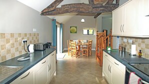 A kitchen with a wooden ceiling beam, a dining area with a table and chairs, and a view of the outside through a window. - The Old Dairy - UKC4698 (Kenilworth)