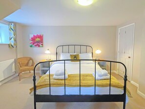 A neatly made bed with white and yellow bedding, two bedside lamps, a wicker chair, a painting of a flower, and a white door. - The Old Dairy - UKC4698 (Kenilworth)