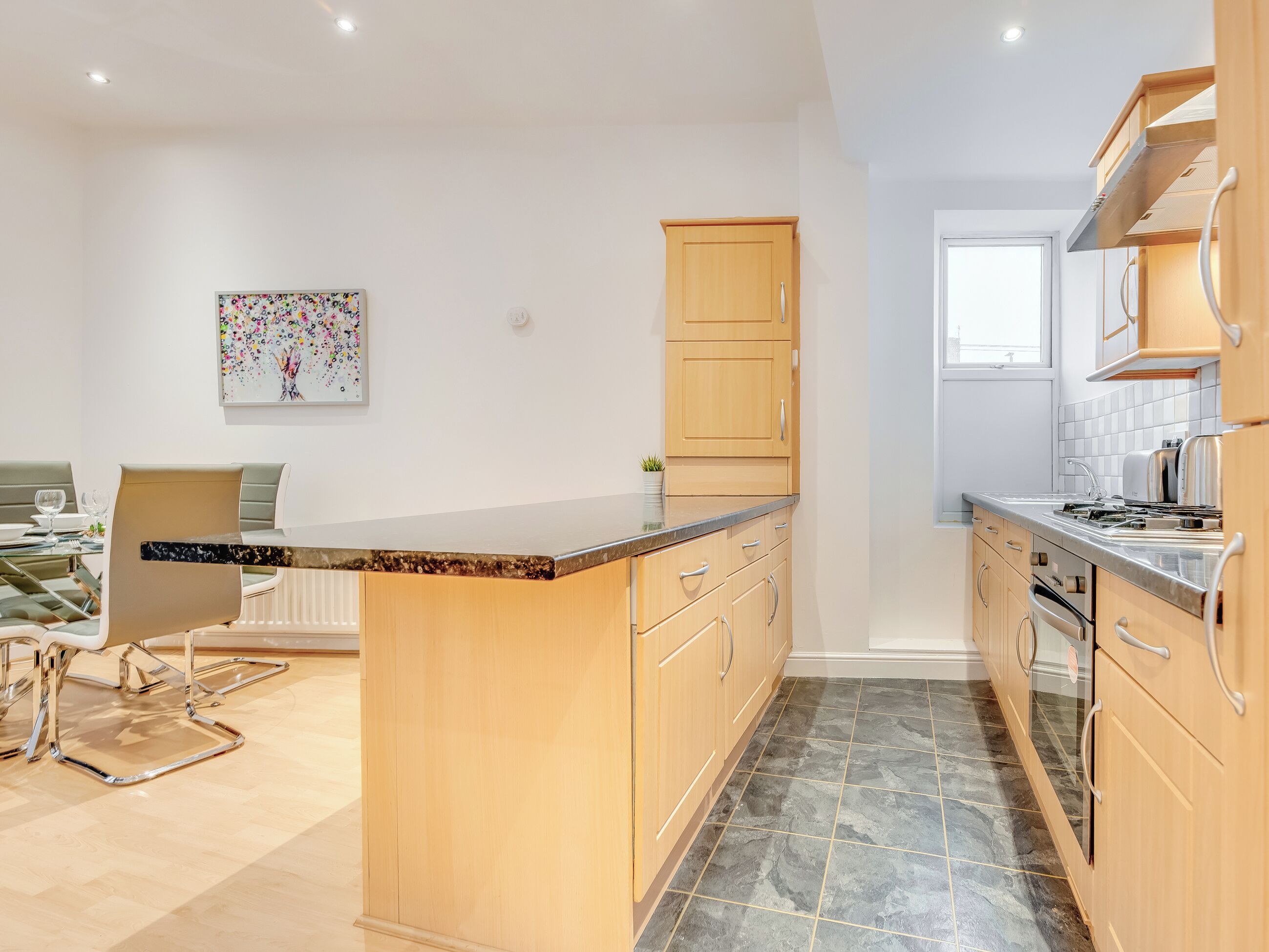 Una cocina moderna con armarios de madera, encimera de mármol y comedor con mesa y sillas de vidrio.