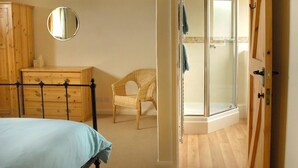 Een slaapkamer met een bed, een houten dressoir, een rieten stoel, een douche met glazen wanden en een deur naar een andere kamer.