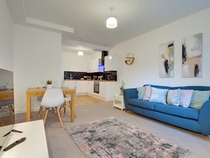 Ein modernes Wohnzimmer mit einem blauen Sofa, einem Essbereich mit Holztisch und einer Küche im Hintergrund.
