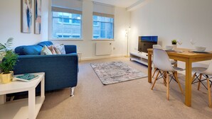 Ein modernes Wohnzimmer mit einem blauen Sofa, einem Esstisch mit Stühlen, einem Fernseher und einer Stehlampe.