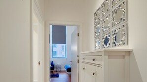 Un couloir au plancher de bois mène à une pièce où se trouvent un lit et une chaise. Un panneau décoratif orne le mur au-dessus d’une armoire.