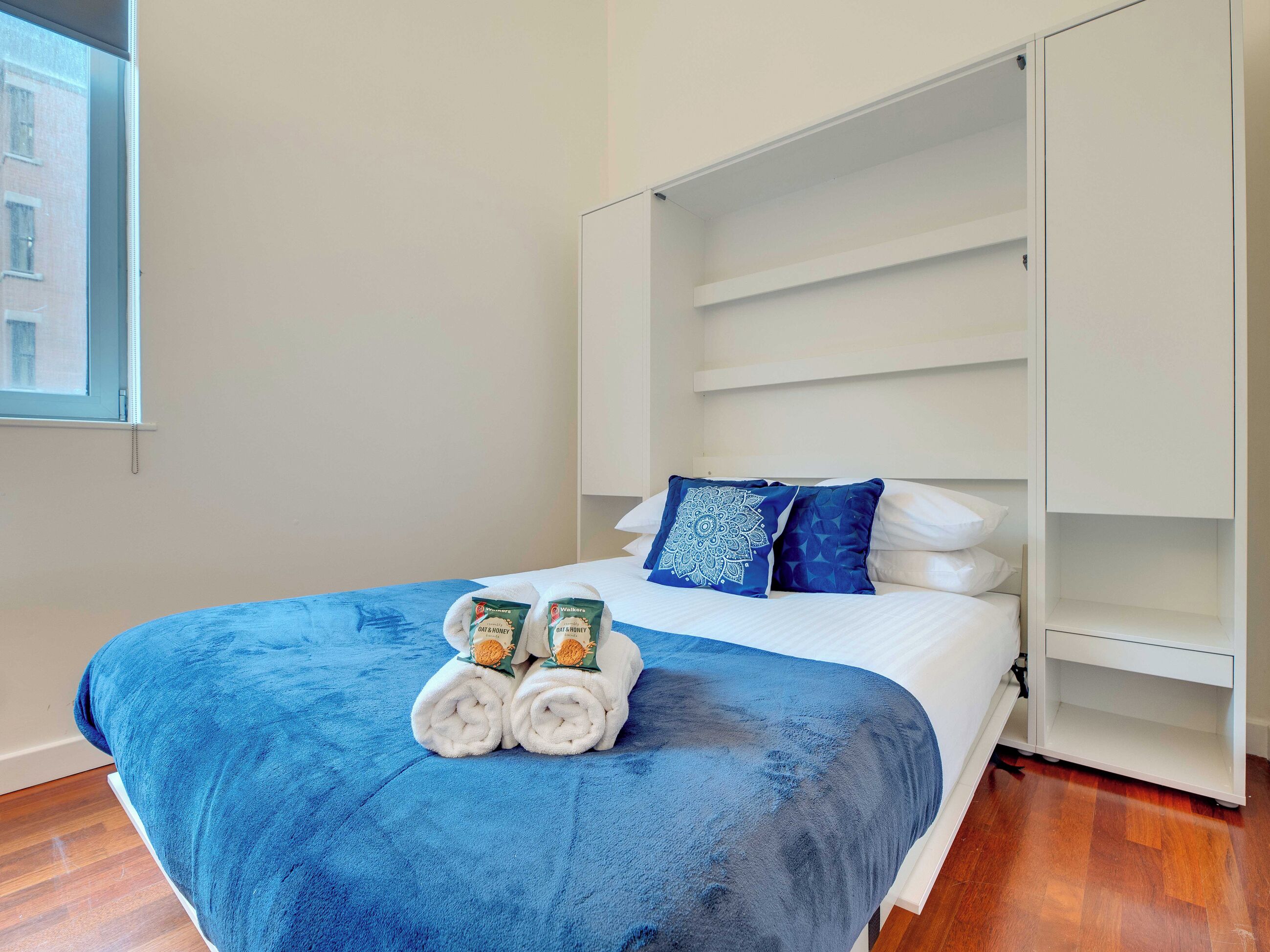 Een slaapkamer met een bed, een blauwe deken, twee opgerolde handdoeken, een wit hoofdeinde en een raam met uitzicht op gebouwen.