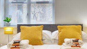 Een keurig opgemaakt bed met witte en mosterdkleurige kussens, een bedlampje en een kleine plant op de vensterbank.