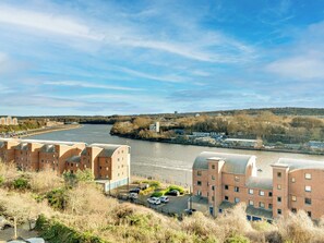 A riverside view with a row of brick buildings, a parking area, and a clear sky. - 2 bedroom accommodation in Newcastle Upon Tyne (Newcastle Upon Tyne)