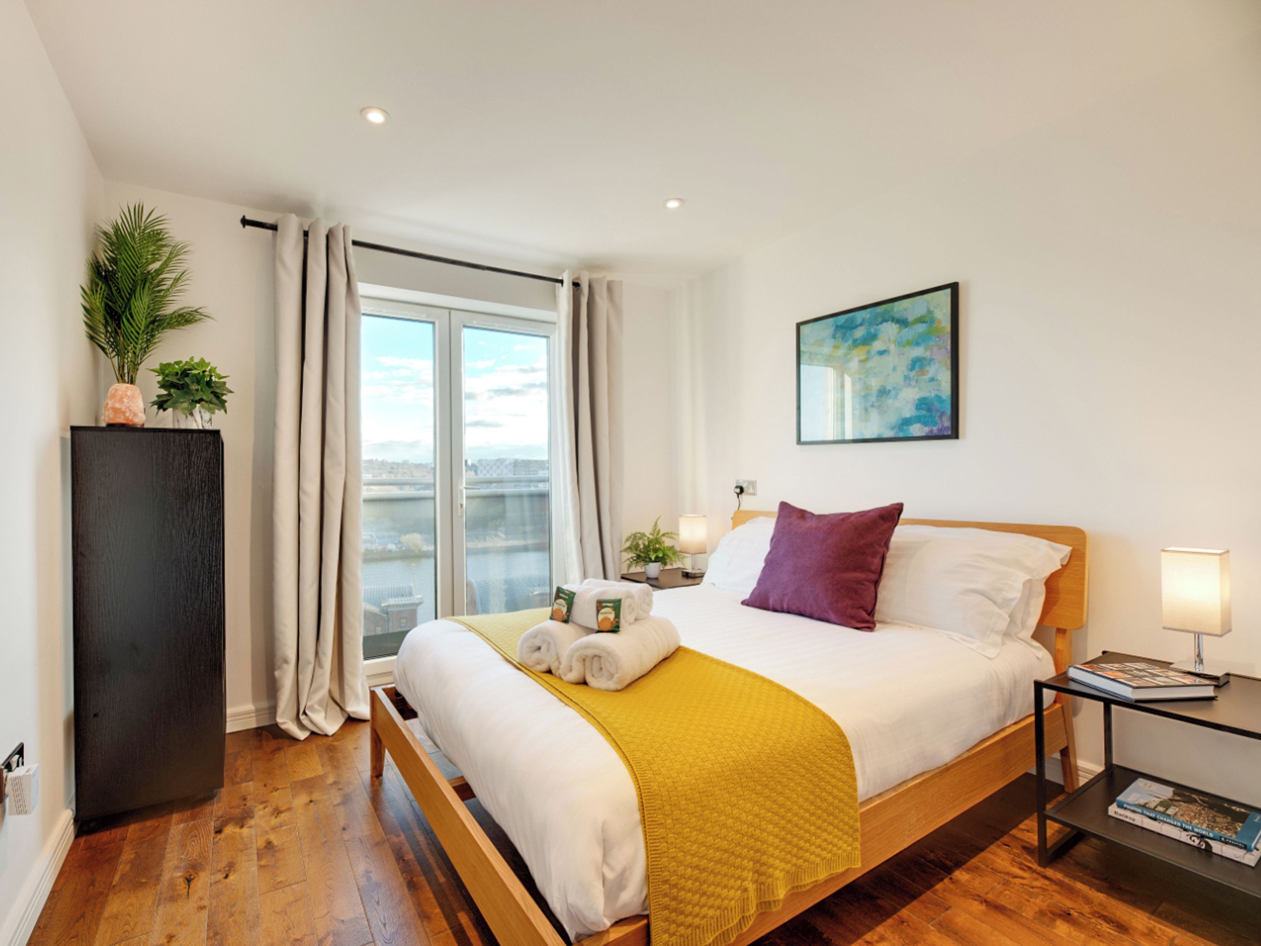 Una habitación de hotel con cama, mesita de noche, ventana con cortinas, una planta y un cuadro en la pared.