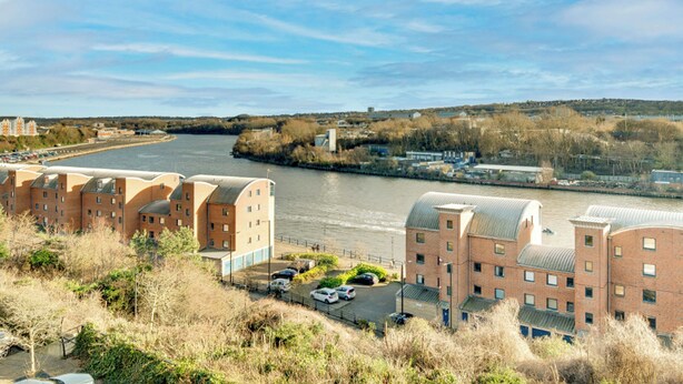A riverside view with a row of brick buildings, a parking area, and a clear sky. - 2 bedroom accommodation in Newcastle Upon Tyne (Newcastle Upon Tyne)