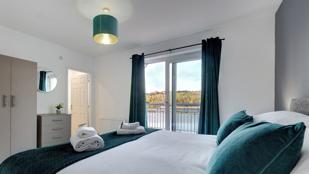 Une chambre moderne avec un grand lit, une commode, un miroir et une vue sur une rivière et des arbres, visible par la fenêtre.