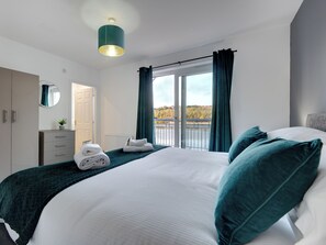A modern bedroom with a large bed, a dresser, a mirror, and a view of a river and trees through a window. - 4 bedroom accommodation in Gateshead (Gateshead)