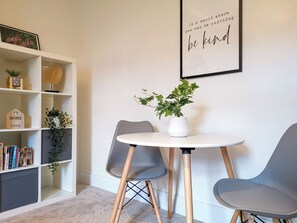 Ein modernes Zimmer mit einem weißen Bücherregal, einem runden Tisch mit zwei Stühlen und einem gerahmten Motivationsspruch an der Wand.