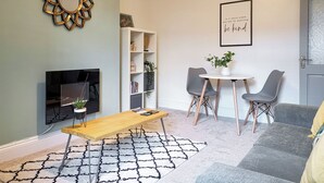Una sala de estar moderna con un televisor de pantalla plana, una mesa de centro de madera, una estantería, dos sillones grises, una pequeña mesa redonda con una planta, y una cita inspiradora enmarcada en la pared.