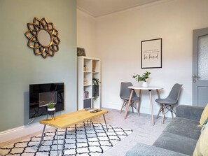 Ein modernes Wohnzimmer mit einem Flachbildfernseher, einem Holztisch, einem Bücherregal, zwei grauen Sesseln, einem kleinen runden Tisch mit einer Pflanze und einem gerahmten, motivierenden Zitat an der Wand.