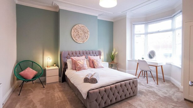 Un dormitorio con cama y cabecera capitonada, una silla verde, un escritorio y un ventanal grande.