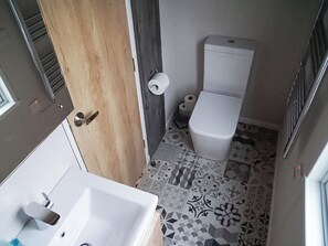 Ein kleines Badezimmer mit weißem Waschbecken, Toilette und Dusche. Der Boden ist mit gemusterten Fliesen ausgelegt, und es gibt eine Holztür.