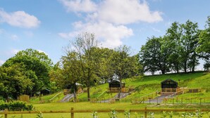Ländliche Landschaft mit Holzhütten auf einer grasbewachsenen Anhöhe, umgeben von Bäumen und einem Zaun.