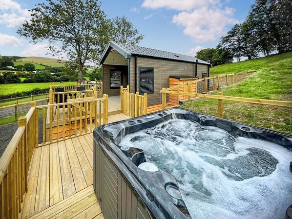 Un bain à remous sur une terrasse en bois, avec vue sur un petit bâtiment et une colline verdoyante en arrière-plan.