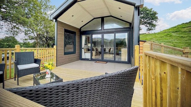 A modern house with a large glass door, a deck with wicker furniture, and a wooden fence. - Hillside Lodge - UKC8191 (Rhayader and the Elan Valley)