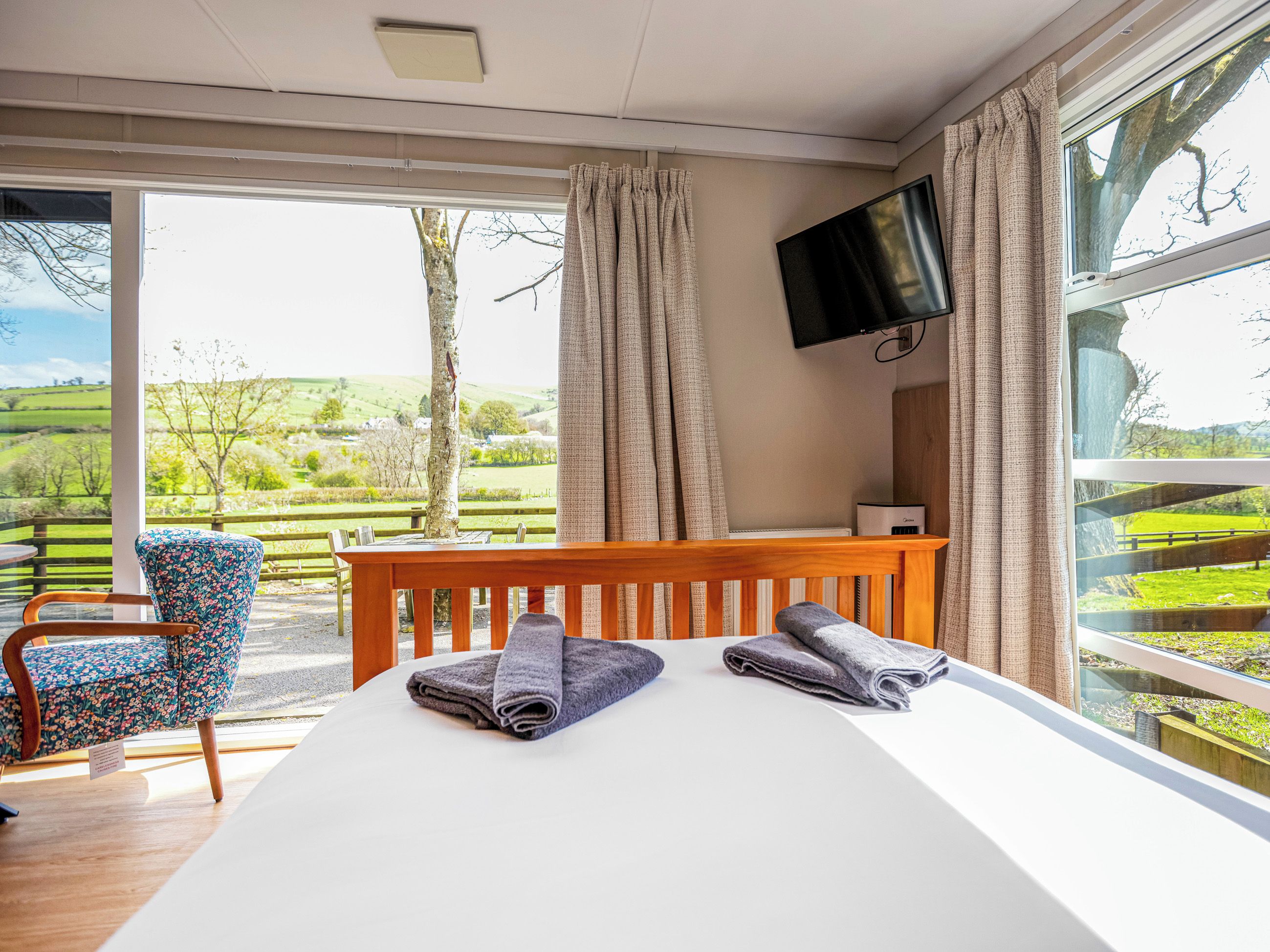 A bedroom with a bed, a TV, a chair, and a view of a green landscape.