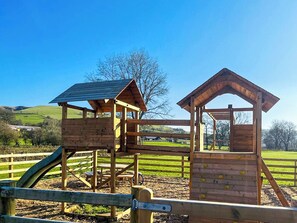 A wooden playground structure with climbing walls and a slide, surrounded by a fence and open fields.