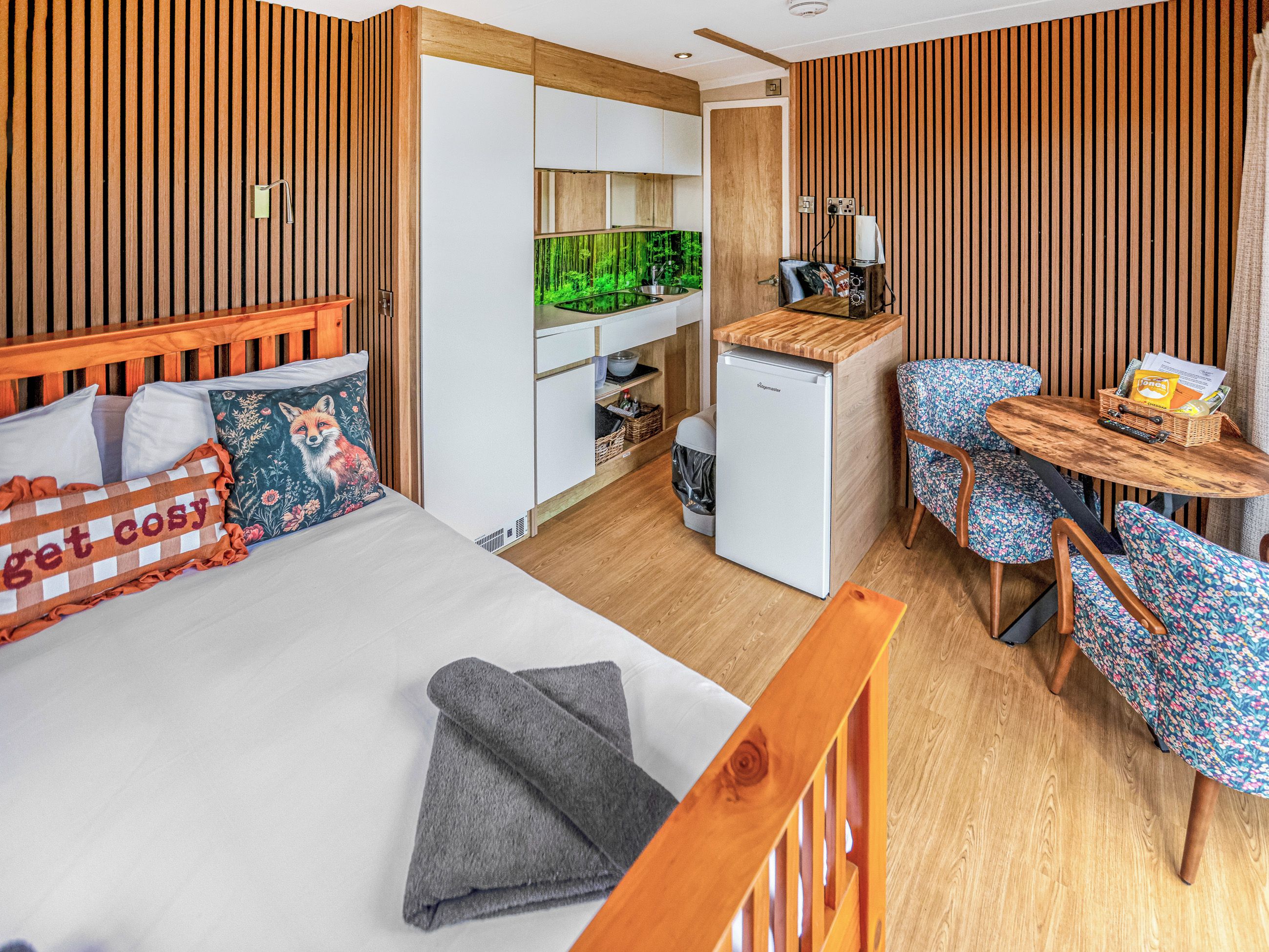 Quarto de hotel compacto com cama, pequeno espaço de cozinha, área de refeições com mesa e cadeiras, e vista para uma floresta verdejante pela janela.