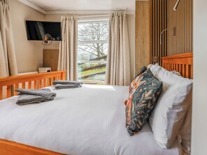 A hotel room with a bed, a TV mounted on the wall, a window with curtains, and a wooden headboard. - The Foxes Den - UKC8196 (Rhayader and the Elan Valley)